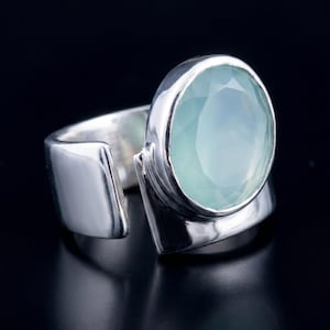May include: A silver ring featuring a large, oval, pale blue gemstone. The ring has a contemporary, open design with a broad band that curves around the finger. The gemstone is faceted, creating a sparkling effect.