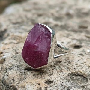 Peut inclure: Bague en argent orn&eacute;e d'une grosse pierre de rubis brute. Le rubis, d'un rouge-ros&eacute; profond, est serti dans une monture en argent avec un anneau fendu. La bague est pr&eacute;sent&eacute;e sur une surface textur&eacute;e de couleur claire.