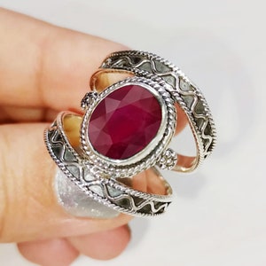 May include: A silver ring with an oval-shaped ruby gemstone set in a decorative silver bezel. The ring has a triple band design with intricate details.