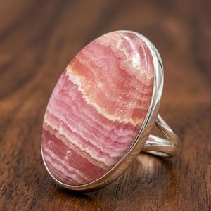 May include: A silver ring featuring a large, oval-shaped gemstone. The gemstone displays a layered pattern of pink and white hues. The ring's band is a simple, polished silver design, complementing the stone's natural beauty.