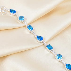 May include: A silver bracelet with multiple blue gemstones in various shapes, including oval, teardrop, and rectangular cuts. The bracelet is displayed on a cream-colored satin fabric.