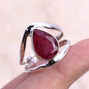 May include: A silver ring featuring a teardrop-shaped ruby gemstone. The ring has a distinctive, open design with two bands that meet at the top.