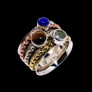 Peut inclure: Une bague multicolore avec une base argentée et plusieurs bandes en cuivre, or et argent. La bague comporte trois pierres précieuses : une bleue, une marron et une verte. La bague est posée sur un fond noir.