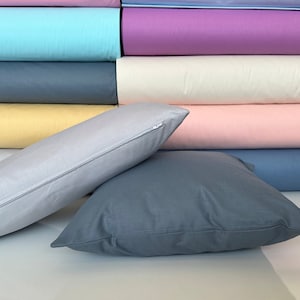 May include: Two pillows, one gray and one light gray, are arranged on a surface with a stack of colorful fabric swatches behind them. The gray pillow has a zipper closure.