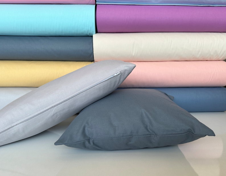 May include: Two pillows, one gray and one light gray, are arranged on a surface with a stack of colorful fabric swatches behind them. The gray pillow has a zipper closure.