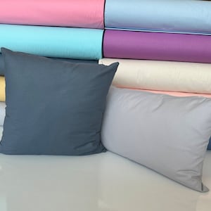 May include: Two decorative pillows, one dark blue and one light gray, are displayed on a white surface. The pillows are in front of a stack of colorful fabric rolls, including pink, light blue, purple, white, yellow, and light pink.