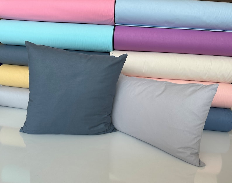 May include: Two decorative pillows, one dark blue and one light gray, are displayed on a white surface. The pillows are in front of a stack of colorful fabric rolls, including pink, light blue, purple, white, yellow, and light pink.