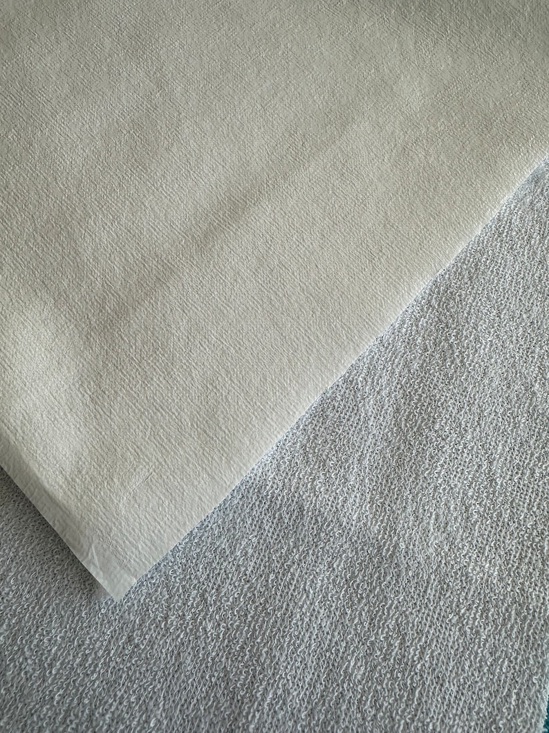 May include: Two pieces of fabric, one white and one gray, are laid on top of each other. The white fabric is smooth and the gray fabric is textured.