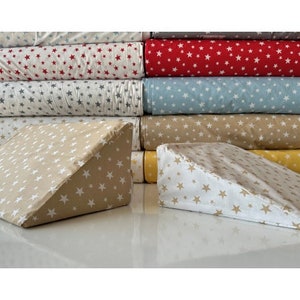 May include: Two beige wedge pillows with a white star pattern. The pillows are stacked on top of a stack of fabric with a white star pattern on a beige background. The fabric is in various colors, including red, blue, and beige.