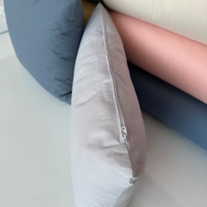 May include: A light gray pillow with a zipper closure. The pillow is lying on a white surface.
