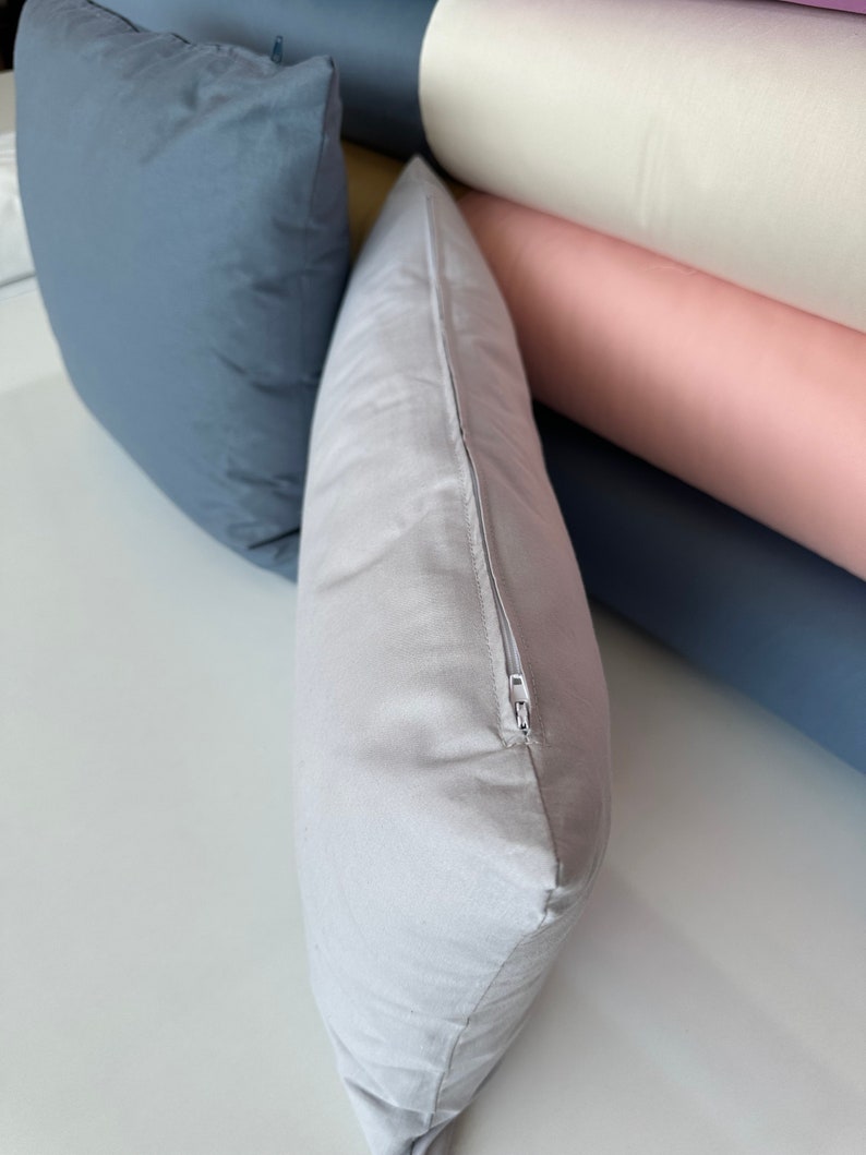 May include: A light gray pillow with a zipper closure. The pillow is lying on a white surface.
