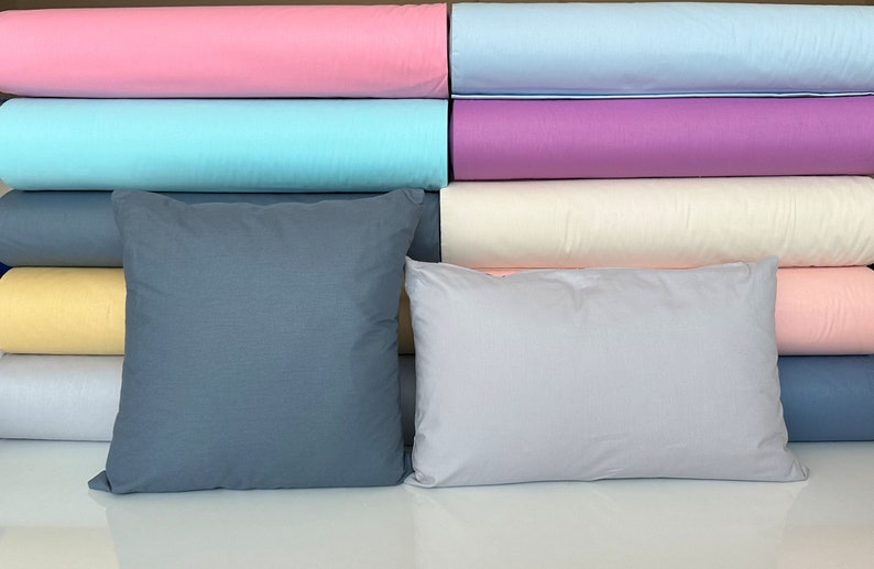 May include: Two pillows, one gray and one light gray, are in front of a stack of fabric rolls in various colors, including pink, light blue, purple, cream, yellow, and light blue.