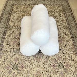 Könnte beinhalten: Drei weiße zylindrische Kissen, die auf einem gemusterten Teppich angeordnet sind. Die Kissen sind aus weichem, weißem Stoff gefertigt und eignen sich perfekt, um jedem Raum Komfort und Stil zu verleihen.