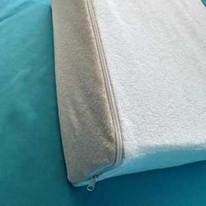 May include: A white, zippered wedge pillow with a soft, textured fabric. The pillow is on a teal surface.