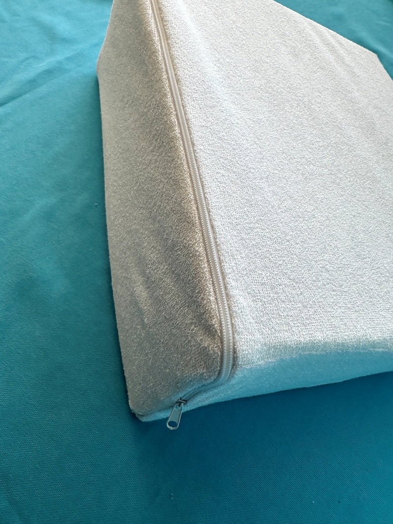 May include: A white, zippered wedge pillow with a soft, textured fabric. The pillow is on a teal surface.