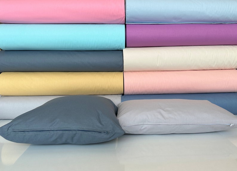 May include: A stack of fabric rolls in various colors, including pink, blue, yellow, and cream. Two pillows are in the foreground, one is gray and the other is light gray.