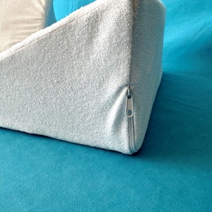 May include: A light blue wedge pillow with a zipper closure. The pillow is made of a soft, textured fabric and is lying on a teal surface.