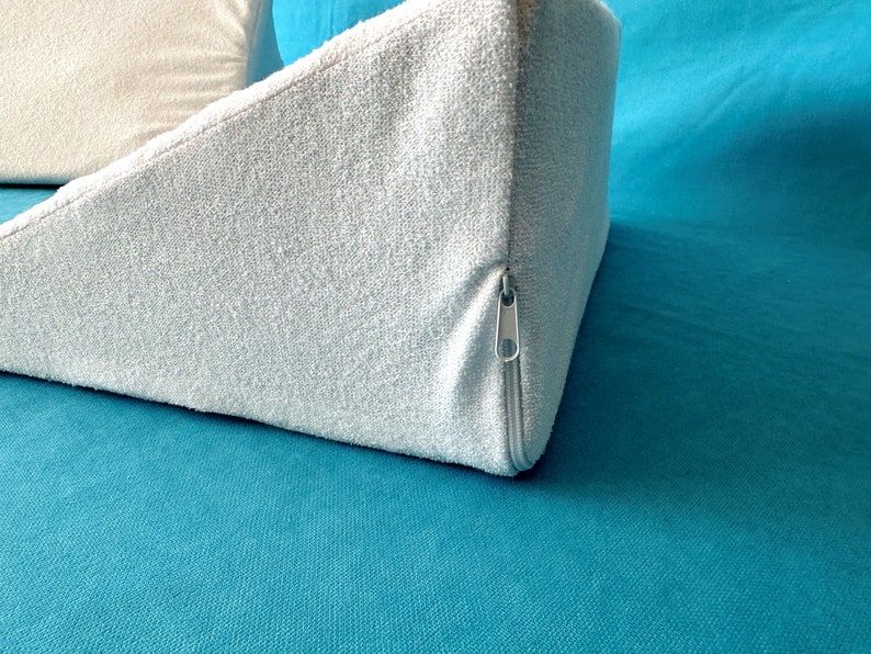 May include: A light blue wedge pillow with a zipper closure. The pillow is made of a soft, textured fabric and is lying on a teal surface.