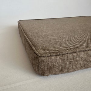 May include: A brown square cushion with a textured fabric cover. The cushion has a thick, stitched border.