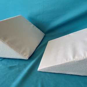 May include: Two white triangular foam wedges on a turquoise blue surface. The wedges are made of a soft, plush material and are designed to provide support and comfort.