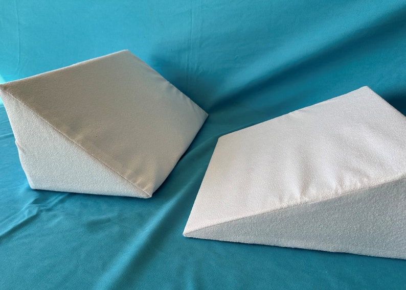 May include: Two white triangular foam wedges on a turquoise blue surface. The wedges are made of a soft, plush material and are designed to provide support and comfort.