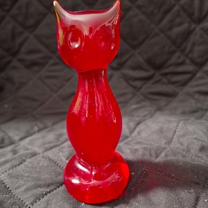 May include: A translucent red glass figurine shaped like a cat with pointed ears and circular eyes. The object features a smooth, rounded body that tapers at the neck and rests on a circular base, reflecting light through its vibrant red material.