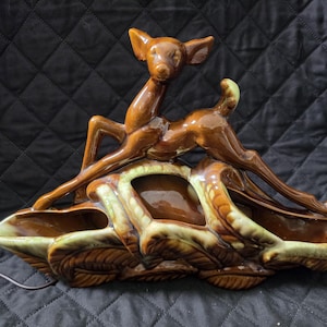 May include: A glossy brown ceramic planter featuring a sculpted deer standing atop a base designed with textured, overlapping leaves. The planter has a smooth, reflective finish with light green highlights along the edges of the leaves and deer.