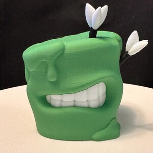 May include: A green 3D printed planter shaped like a smiling face with white teeth and two white darts sticking out of the top.