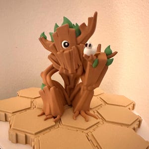 Puede incluir: Una figura de árbol de color marrón y verde con un solo ojo y un pequeño pájaro blanco posado en su brazo. La figura está de pie sobre una base hexagonal.