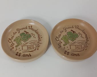 Könnte beinhalten: Zwei beige Keramikplatten mit einer braunen Illustration eines Gebäudes und Trauben. Auf den Tellern steht der Text "Chai du grand Vaudamière" und "25 ans". Die Teller sind rund und haben einen leicht erhöhten Rand.