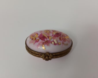 Könnte beinhalten: Eine kleine ovale Schmuckschatulle mit einem floralen Design in Rosa und Gold auf weißem Hintergrund. Die Schachtel hat einen goldenen Metallrand und einen Klappdeckel.