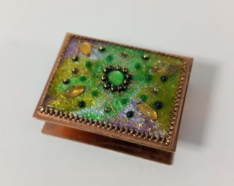 Puede incluir: Una pequeña caja decorativa cuadrada, verde y dorada, con una joya verde en el centro. La caja tiene un marco de cobre y está decorada con un patrón floral.