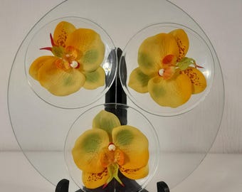 May include: A clear circular glass display stand featuring three integrated smaller glass circles, each adorned with a single artificial yellow and green orchid flower. The entire glass structure is supported by a black plastic tripod stand.