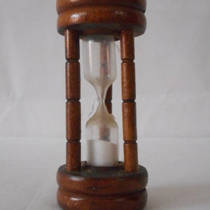 Puede incluir: Un reloj de arena de madera con acabado marrón oscuro. El reloj de arena tiene un centro de vidrio transparente lleno de arena blanca. El marco de madera se compone de dos bases circulares conectadas por cuatro soportes verticales.