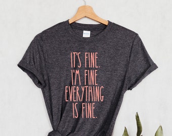 以下が含まれることがあります： ピンクの文字で「It's fine. I'm fine. Everything is fine.」とプリントされたダークグレーのTシャツです。