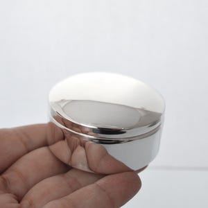 May include: A small, round, polished silver-coloured container with a smooth, reflective surface and a lid. The object has a cylindrical shape with a flat top and a slightly recessed base, featuring a clean, metallic finish that reflects light.