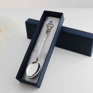 May include: A silver spoon with an ornate design and a blue gemstone on the handle. The spoon is in a blue velvet box.