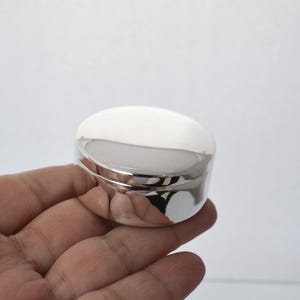 Könnte beinhalten: Ein kleiner, runder, silberfarbener Behälter mit Deckel. Der Behälter ist hochreflektierend, mit einer glatten, polierten Oberfläche. Der Deckel ist leicht gewölbt, und der Behälter scheint aus Metall zu sein. Der Hintergrund ist weiß.