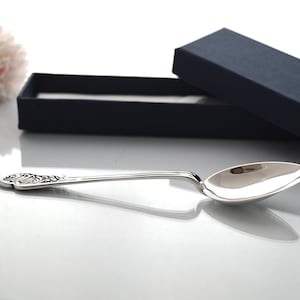 Puede incluir: Una cuchara plateada con un mango adornado y un cuenco pulido, presentada en una caja azul oscuro. Una flor rosa está en el fondo. La cuchara es probablemente de plata de ley.