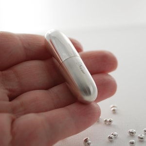 May include: A smooth, cylindrical silver-toned metal pill case with a rounded top and bottom. The surface features the word Name engraved in a cursive script font. The object is small enough to be held in the palm of a hand, reflecting light.