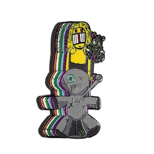 May include: An enamel pin with a distinctive cartoon design. It depicts a yellow-haired character with an open mouth, a grey voodoo doll, and a green teddy bear. The design incorporates a rainbow colour scheme and is set against a white background.