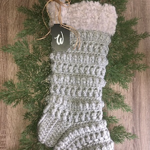 May include: A gray, handmade Christmas stocking with a fluffy, light gray cuff. The stocking is crocheted with a textured stitch and has a loop for hanging. A black tag with the letter "W" is attached with twine. The stocking is displayed on a wooden surface with evergreen branches.