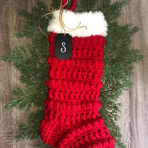 May include: Red crocheted Christmas stocking with a white faux fur cuff and a black tag with the letter "S" on it.