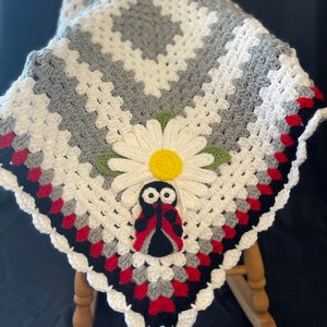 May include: A crocheted baby blanket with a white, gray, red, and black color scheme. The blanket features a daisy flower and a ladybug applique.