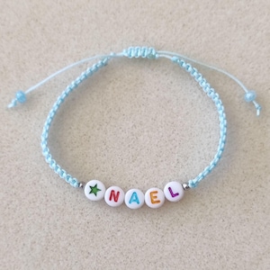 Op de afbeelding: Lichtblauw macramé armband met witte letterkralen met de tekst "NAEL". De armband heeft een groene ster kraal en verstelbare schuifknopen. Een gepersonaliseerd accessoire.