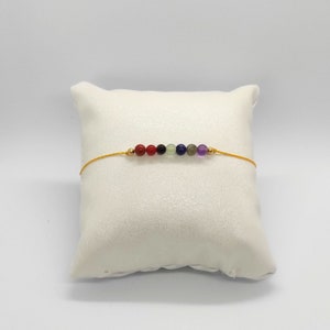 Può includere: Un braccialetto di corda gialla con sette perline rotonde di pietre preziose in un motivo arcobaleno. Le perline sono rosse, nere, verdi, blu, viola e trasparenti.