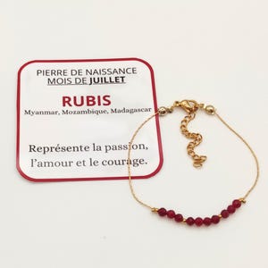Peut inclure: Un bracelet doré orné de petites perles rouges facettées. Il est présenté à côté d'une carte indiquant « RUBIS » et « MOIS DE JUILLET ». La carte précise également l'origine des rubis : Myanmar, Mozambique et Madagascar.