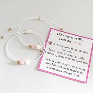 Peut inclure: Deux bracelets en fil blanc avec des perles roses et blanches et des détails dorés. Une étiquette rose avec du texte français est attachée, indiquant "Duo mère et fille - Lien du coeur". Les bracelets sont conçus pour rappeler un lien spécial.