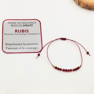 Può includere: Braccialetto in corda rossa con piccole perle di pietre preziose rosse e dettagli dorati. Il braccialetto è legato con un nodo scorrevole. Un biglietto con il testo "PIERRE DE NAISSANCE MOIS DE JUILLET RUBIS Myanmar, Mozambique, Madagascar Représente la passion, l'amour et le courage." è sullo sfondo.