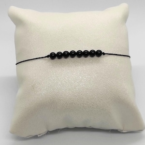Puede incluir: Una pulsera de cuentas negras con siete cuentas redondas y pulidas ensartadas en un fino cordón negro. La pulsera se muestra sobre un cojín blanco texturizado, creando un contraste entre las cuentas oscuras y el fondo claro.